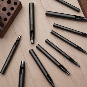May include: An assortment of dark gray leathercraft tools, including hole punches and a wooden tool holder. The tools are arranged on a light wood surface, showcasing their design and functionality. The tools are labeled with the brand name.
