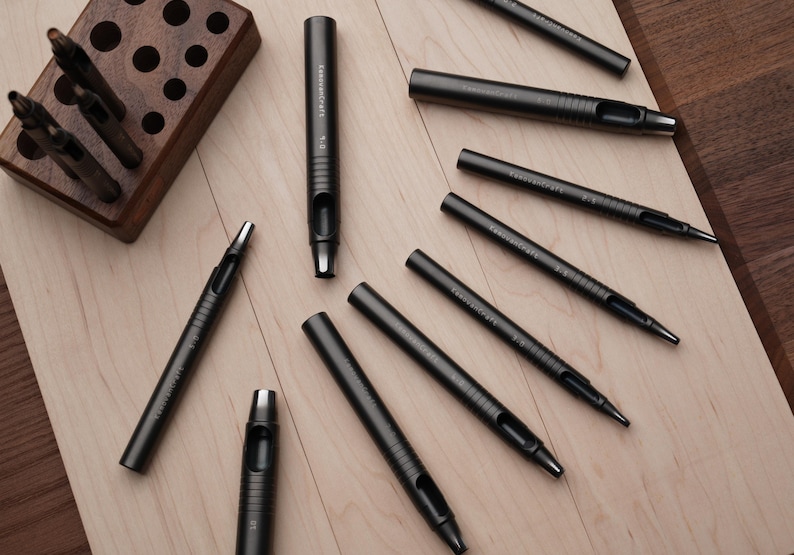 May include: An assortment of dark gray leathercraft tools, including hole punches and a wooden tool holder. The tools are arranged on a light wood surface, showcasing their design and functionality. The tools are labeled with the brand name.
