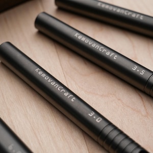 May include: Close-up of several dark gray metal tools with the text "KemovanCraft" and numbers indicating sizes. The tools are cylindrical with ridged grips and are arranged on a light wood surface.