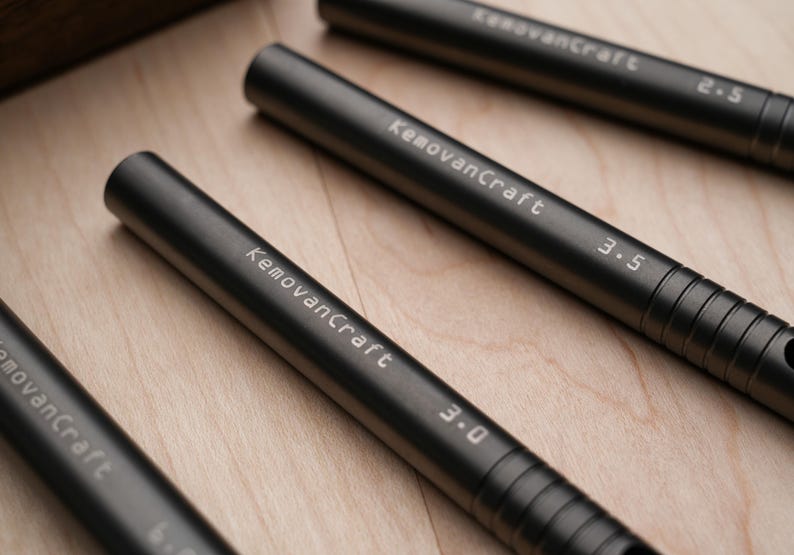 May include: Close-up of several dark gray metal tools with the text "KemovanCraft" and numbers indicating sizes. The tools are cylindrical with ridged grips and are arranged on a light wood surface.