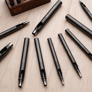 May include: A collection of dark gray leathercraft tools, including various sizes of hole punches and a wooden storage box. The tools are arranged on a light wood surface, showcasing their metallic finish and precision design.