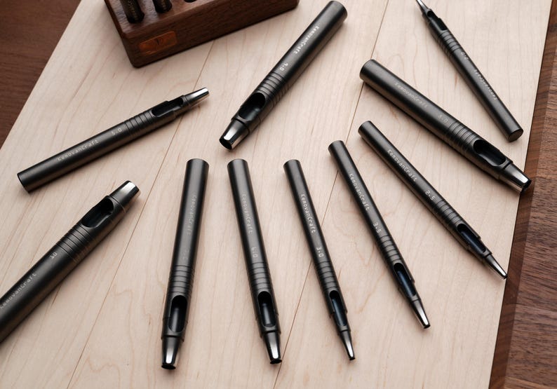 May include: A collection of dark gray leathercraft tools, including various sizes of hole punches and a wooden storage box. The tools are arranged on a light wood surface, showcasing their metallic finish and precision design.