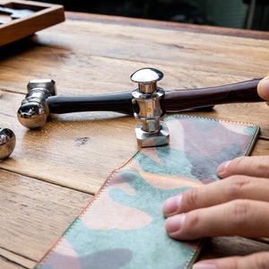 Puede incluir: Dos martillos de metal con mangos de madera se utilizan para presionar un trozo de cuero con estampado de camuflaje. El cuero está siendo sujetado por una mano.