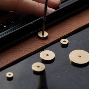 May include: A set of six brass rings of varying sizes, with a ruler and a tool used to punch holes in leather. The rings are arranged on a brown leather surface.