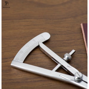 May include: A silver metal leather measuring tool with a protractor and a screw adjustment. The tool is on a wooden surface.