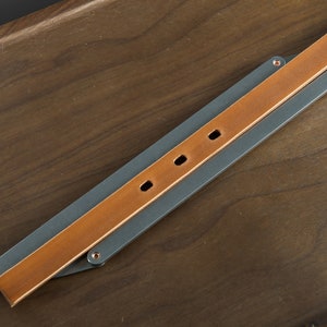 May include: A brown leather strap with three copper rivets is attached to a gray metal strap. The straps are folded in half and laid on a wooden surface.