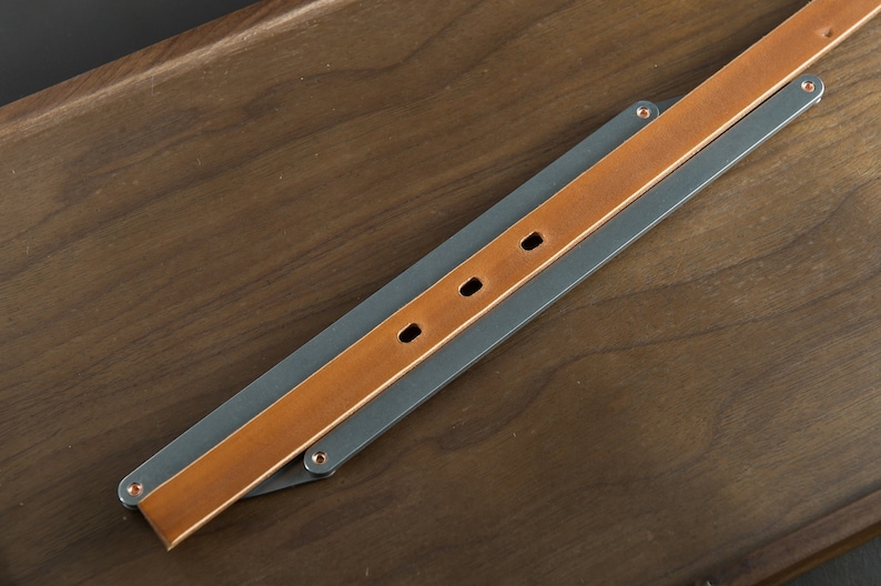 May include: A brown leather strap with three copper rivets is attached to a gray metal strap. The straps are folded in half and laid on a wooden surface.