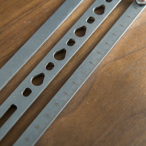May include: A silver metal ruler with a series of cut-out shapes along the top edge. The ruler has markings in inches.