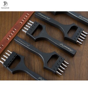 May include: Black leatherworking tools with multiple prongs, arranged on a wooden surface. The tools have the text "KemoranCraft" engraved on them. A brown leather strap with punched holes is also visible.