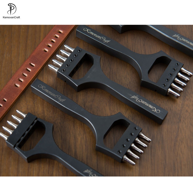 May include: Black leatherworking tools with multiple prongs, arranged on a wooden surface. The tools have the text "KemoranCraft" engraved on them. A brown leather strap with punched holes is also visible.