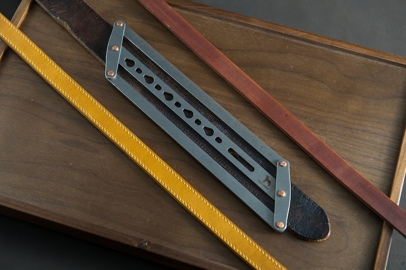 May include: A metal tool with a leather strap and two other leather straps in yellow and brown. The tool has a series of holes and a metal logo with the text "The Leatherworker".
