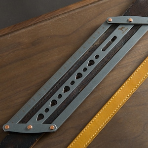 May include: A metal ruler with a leather strap and copper rivets. The ruler has a series of holes and markings along its length.