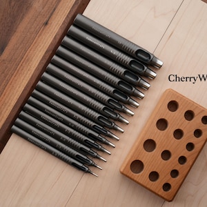 May include: A set of dark gray metal hole punches with silver tips, arranged diagonally on a light wood surface. A cherry wood block with various sized holes sits next to the punches. The text "CherryWood" is visible.