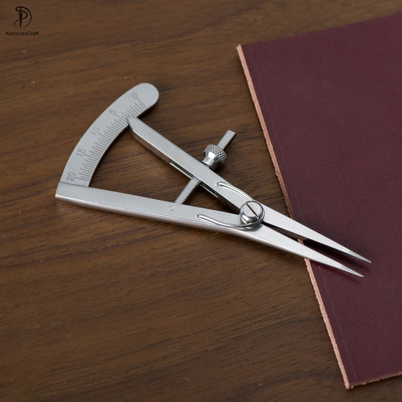 May include: A silver leather crafting tool with a protractor and adjustable arms. The tool is on a brown wooden surface.