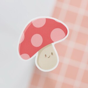 May include: A cute mushroom sticker. The mushroom has a red cap with light pink spots and a beige stem with a smiling face. The sticker is outlined in white, with a cartoon style.