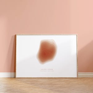May include: A framed print featuring a topographic map design of Cairn Gorm in the Scottish Highlands. The artwork showcases a layered, abstract representation in shades of orange and beige against a white background. The print is displayed in a light wood frame.