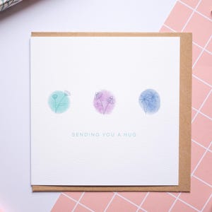 May include: A white card with a brown envelope. The card features three watercolour circles containing blue, purple, and green flowers. The text "Sending you a hug" is printed below the circles.