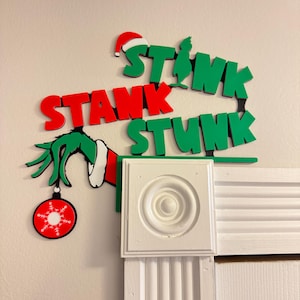 May include: Green and red Christmas decoration with the words "Stink Stank Stunk" and a green Grinch hand holding a red ornament with a snowflake.