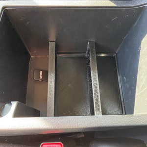 May include: A black plastic car center console with three compartments. The console has a red button labeled "PRESS" and a small black device labeled "AUX IN".