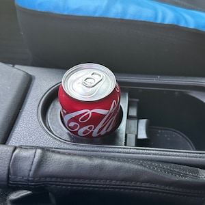 May include: A red aluminum can with the word "Coca-Cola" printed on it, sitting in a black cup holder in a car.