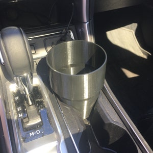 May include: A black plastic cup holder for a car. The cup holder is designed to fit in the center console of a car, near the gear shift.