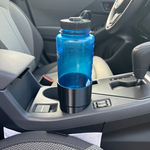 May include: A blue water bottle with a black lid and a black carrying strap is in a cup holder in a car. The cup holder is located in the center console of the car.