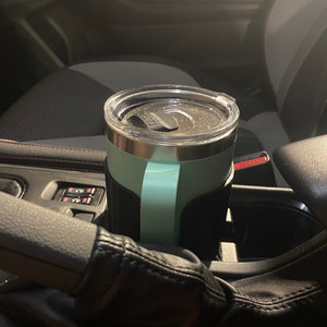 May include: A teal travel mug with a black sleeve is in a car cup holder. The mug has a clear lid and a stainless steel body.