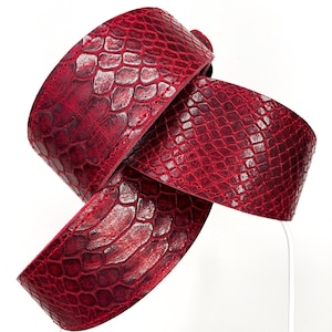 Pode incluir: Um cinto vermelho com um padrão de pele de cobra. O cinto é feito de um material texturizado e é exibido em um fundo branco. O cinto está dobrado sobre si mesmo.