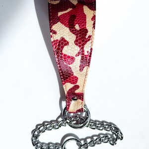 Peut inclure: Une sangle en cuir à motif camouflage rouge et beige avec une chaîne et des anneaux argentés. La sangle est fixée à une chaîne avec deux anneaux argentés et un plus grand anneau à l'extrémité.