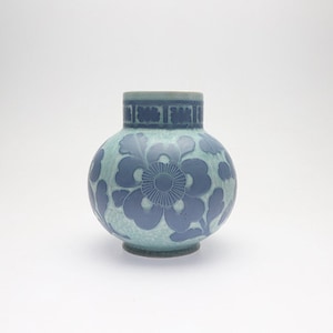 Puede incluir: Un jarrón redondo de cerámica con una base de color verde azulado claro y un patrón floral azul. El jarrón tiene un cuello corto con una banda decorativa. El diseño floral es una característica destacada, que añade elegancia.