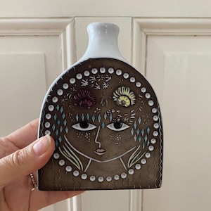 May include: A brown ceramic vase with a white neck, decorated with a stylized face design. The face has large eyes, a small mouth, and is decorated with floral and geometric patterns. The vase is held by a hand.