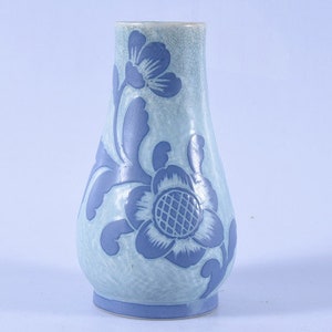 Puede incluir: Un jarrón de cerámica azul claro con un diseño floral en un tono azul más oscuro. El jarrón tiene forma bulbosa y una abertura ligeramente acampanada. El patrón floral incluye una flor grande y hojas. El jarrón mide unos 15 cm de alto.