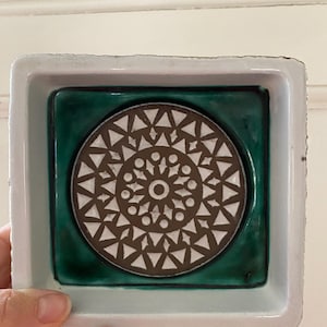 May include: A square ceramic dish with a white rim and a dark green interior. The centre features a circular design with a brown and white geometric pattern. The dish is held by a hand.