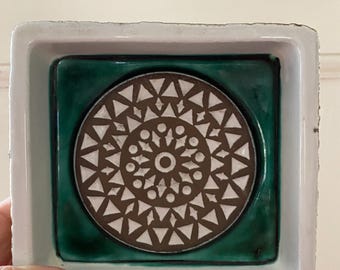 May include: A square ceramic dish with a white rim and a dark green interior. The centre features a circular design with a brown and white geometric pattern. The dish is held by a hand.