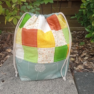 Könnte beinhalten: Ein Patchwork-Beutel mit Kordelzug und einer Mischung aus bunten Quadraten in Orange, Gelb, Grün und Weiß. Der untere Teil ist helltürkis mit einem weißen Blumenmuster. Der Beutel hat einen Kordelzugverschluss und steht auf einer Steinoberfläche.