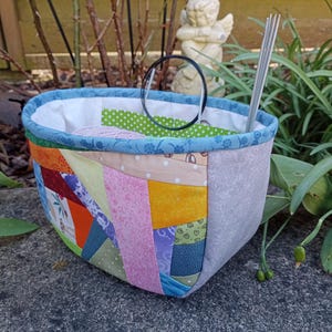 K&ouml;nnte beinhalten: Ein Stoffkorb mit einer Patchwork-Au&szlig;enseite in verschiedenen bunten Mustern, darunter Orange, Gelb, Rosa und Gr&uuml;n. Der Korb hat einen blauen floralen Rand, ein wei&szlig;es Innenfutter und enth&auml;lt einen schwarzen kreisf&ouml;rmigen Ring sowie silberne Stricknadeln.