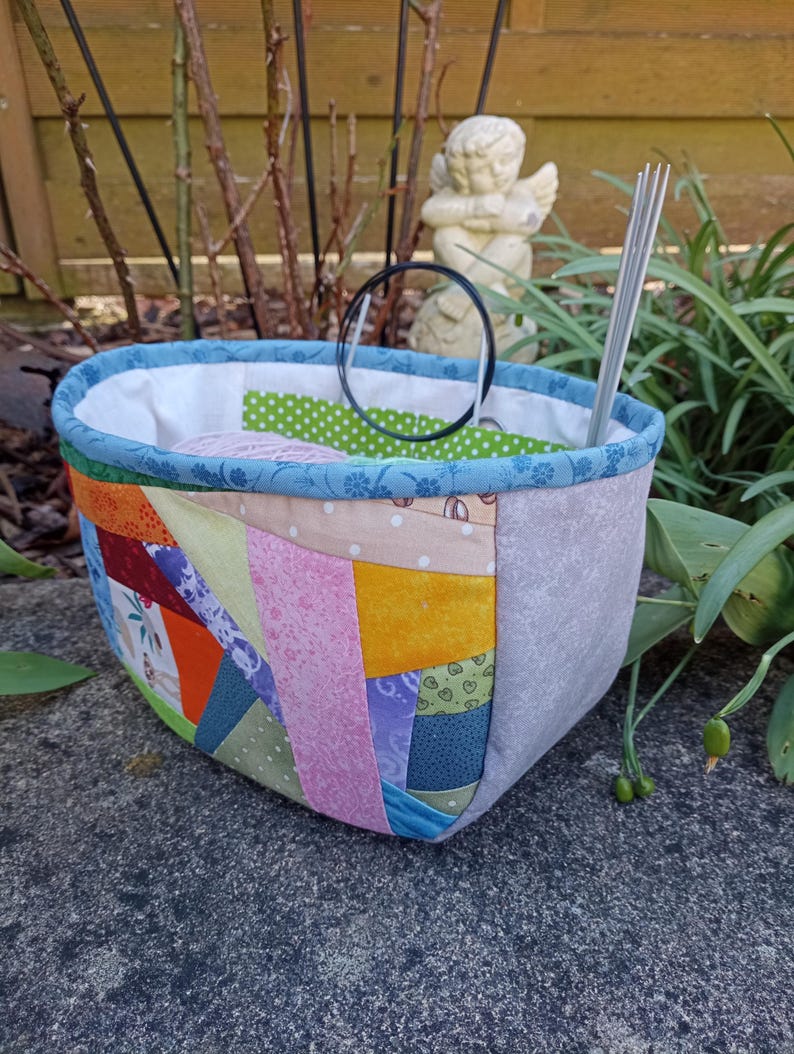 K&ouml;nnte beinhalten: Ein Stoffkorb mit einer Patchwork-Au&szlig;enseite in verschiedenen bunten Mustern, darunter Orange, Gelb, Rosa und Gr&uuml;n. Der Korb hat einen blauen floralen Rand, ein wei&szlig;es Innenfutter und enth&auml;lt einen schwarzen kreisf&ouml;rmigen Ring sowie silberne Stricknadeln.