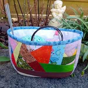 Könnte beinhalten: Ein Stoffkorb mit einer Patchwork-Außenseite in verschiedenen bunten Mustern, darunter Orange, Grün, Rot und Blau. Der Korb hat einen blauen Blumenbesatz, ein weißes Innenfutter und enthält Stricknadeln sowie Wollknäuel.