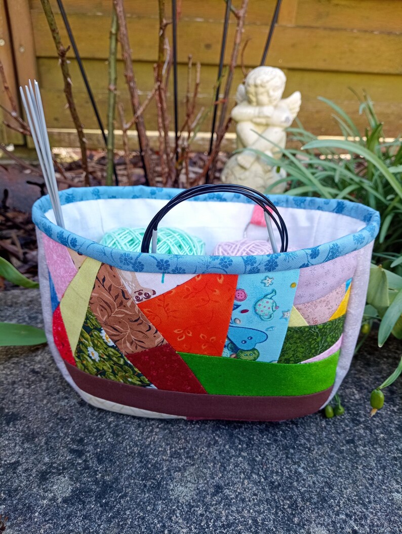 K&ouml;nnte beinhalten: Ein Stoffkorb mit einer Patchwork-Au&szlig;enseite in verschiedenen bunten Mustern, darunter Orange, Gr&uuml;n, Rot und Blau. Der Korb hat einen blauen Blumenbesatz, ein wei&szlig;es Innenfutter und enth&auml;lt Stricknadeln sowie Wollkn&auml;uel.