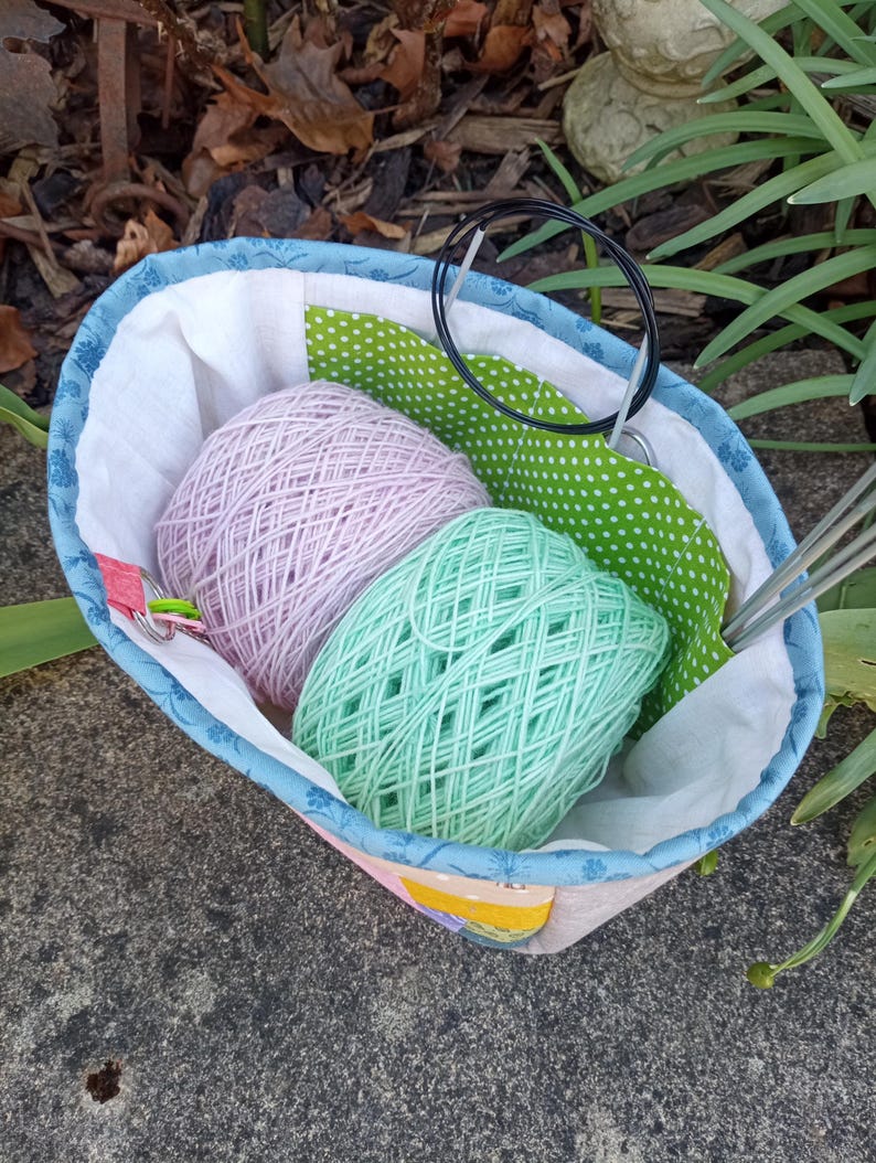 K&ouml;nnte beinhalten: Eine Stofftasche f&uuml;r Handarbeitsprojekte mit blauem Blumenmuster am Rand, die ein Kn&auml;uel hellrosa Garn und ein Kn&auml;uel mintgr&uuml;nes Garn enth&auml;lt. Das Innere ist mit wei&szlig;em Stoff ausgekleidet und hat ein gr&uuml;n-wei&szlig; gepunktetes Paneel sowie Rundstricknadeln.