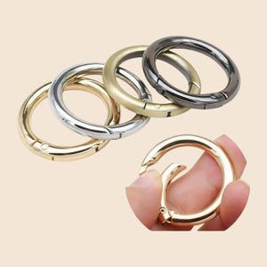 May include: A collection of five circular metal spring gate rings in various finishes including gold, silver, brass, and gunmetal. Each ring features a hinged mechanism that opens and closes, allowing for attachment to bags, keys, or straps.