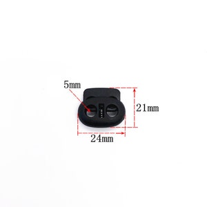 May include: Black plastic toggle buckle with two holes. The buckle measures 24 mm wide, 21 mm tall, and 5 mm thick.