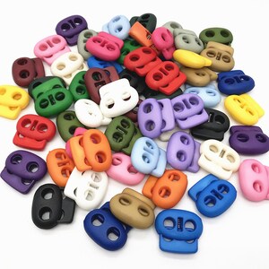 May include: A large assortment of colorful plastic toggle clasps. The clasps are oval shaped with two holes and a spring mechanism. The colors include red, orange, yellow, green, blue, purple, pink, brown, black, and white.
