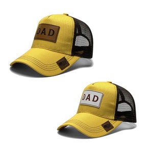 以下が含まれることがあります： 黒いメッシュのバックパネルが付いた黄色のベースボールキャップが2つ。両方のキャップには、赤い文字で「DAD」と書かれた茶色の革のパッチが付いています。