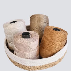 Puede incluir: Cuatro bobinas de cordón en tonos variados de beige, bronceado y marrón están dispuestas dentro de una cesta redonda tejida. Cada bobina presenta un núcleo central de plástico negro y un hilo sintético texturizado con un patrón trenzado.