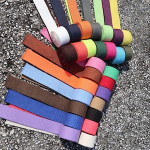 May include: Assortment of colorful cotton webbing straps in various colors, including white, black, brown, orange, red, green, blue, purple, and yellow. The straps are rolled up and arranged in a fan shape.