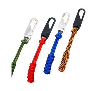 Könnte beinhalten: Vier Reißverschlussanhänger mit geknoteten Paracord-Schlaufen in Olivgrün, Rot, einfarbigem Blau sowie einem schwarz-orangen Zickzackmuster. Jede Schnur ist an einem Karabinerhaken aus Metall befestigt, zwei in Silber und zwei in mattem Schwarz.