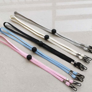 Può includere: Cinque lacci per maschera facciale di diversi colori con tappi regolabili neri. I lacci sono rosa, azzurro, nero, bianco e grigio.