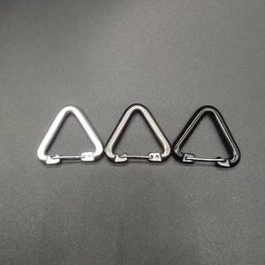 May include: Three triangular carabiners arranged in a row. The first is silver, the second is gunmetal grey, and the third is black. Each carabiner features a spring-loaded gate mechanism that allows for opening and closing the triangular frame.