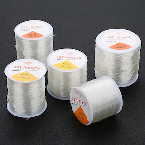 Könnte beinhalten: Vier Spulen mit klarem elastischem Garn zum Schmuckmachen. Jede Spule ist mit "Crystal Tec" beschriftet und hat eine Angabe in Metern.